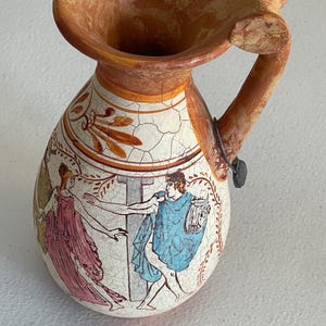 May include: A small, antique-style ceramic pitcher with a wide, flared rim and a handle. The pitcher features a painted scene of figures in classical attire, with a cracked, aged appearance. The rim and handle are a reddish-brown color.