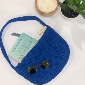 May include: A royal blue, crocheted shoulder bag with a curved shape. The bag contains a book, sunglasses, and a small container of Aveeno. A candle and a plant are in the background.