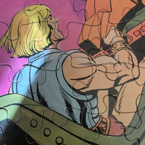 May include: A close-up of a comic book character with blond hair, wearing a blue shirt and brown pants. The character is holding onto a large, orange object with a brown strap.