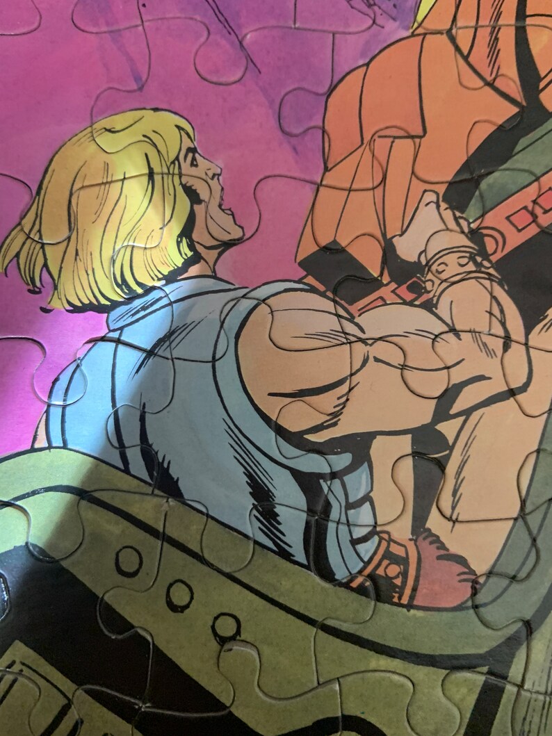 May include: A close-up of a comic book character with blond hair, wearing a blue shirt and brown pants. The character is holding onto a large, orange object with a brown strap.