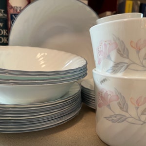 Pode incluir: Um conjunto de pratos de cerâmica branca com borda azul e padrão floral. O conjunto inclui tigelas, pratos e canecas.