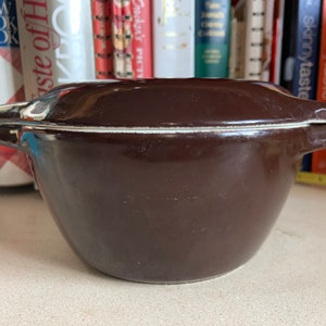 May include: A dark brown, ceramic casserole dish with a lid. The dish has a rounded shape with two handles and a white rim. The lid has a similar shape with two handles. The dish is likely used for cooking and serving food.