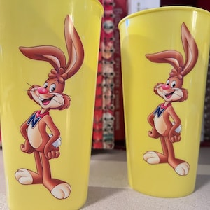 Puede incluir: Dos vasos de plástico amarillos con un conejo de dibujos animados que lleva una camisa marrón y una letra N azul. El conejo sonríe y tiene las manos en las caderas.