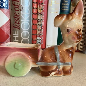 Puede incluir: Una maceta de cerámica con forma de burro tirando de un carro. El burro es marrón y beige con un arnés gris. El carro es rosa y verde. El objeto es decorativo.