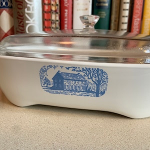 May include: White casserole dish with a clear glass lid. The dish features a blue illustration of a house and trees. The lid has a clear glass knob. The dish is sitting on a light gray surface.