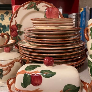 Puede incluir: Un juego de vajilla de cerámica vintage con un motivo de manzana. El juego incluye una mantequillera, un azucarero, una jarra de crema y varios platos. La vajilla está decorada con ramas marrones, hojas verdes y manzanas rojas.