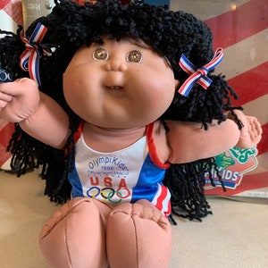 Könnte beinhalten: Eine Cabbage Patch Kid Puppe, die einen blau-weiß-roten Badeanzug mit dem Text "OlympiKids 1996 Official Team Mascot USA" und den olympischen Ringen trägt. Die Puppe hat schwarzes Garnhaar mit rot-weiß-blauen Schleifen.