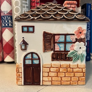 Puede incluir: Una jarra de galletas de cerámica con forma de casita con techo marrón, paredes blancas y fachada de ladrillo. La casa tiene una puerta, dos ventanas y una jardinera con flores.