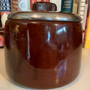 Op de afbeelding: Een bruine keramische pot met een zilveren rand en deksel. De pot heeft twee kleine handvatten aan de zijkanten en een kleine knop op het deksel. De pot is rond en heeft een glanzende afwerking.