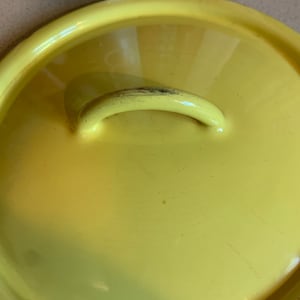 May include: A bright yellow lid with a curved handle. The lid is round and appears to be made of metal or enamel. The handle is slightly worn. The lid is likely for a pot or pan.
