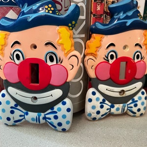 Puede incluir: Dos tapas de interruptor de luz con un rostro de payaso con un sombrero azul, una nariz roja y una pajarita azul y blanca con lunares. Los rostros de payaso están pintados sobre un fondo blanco.