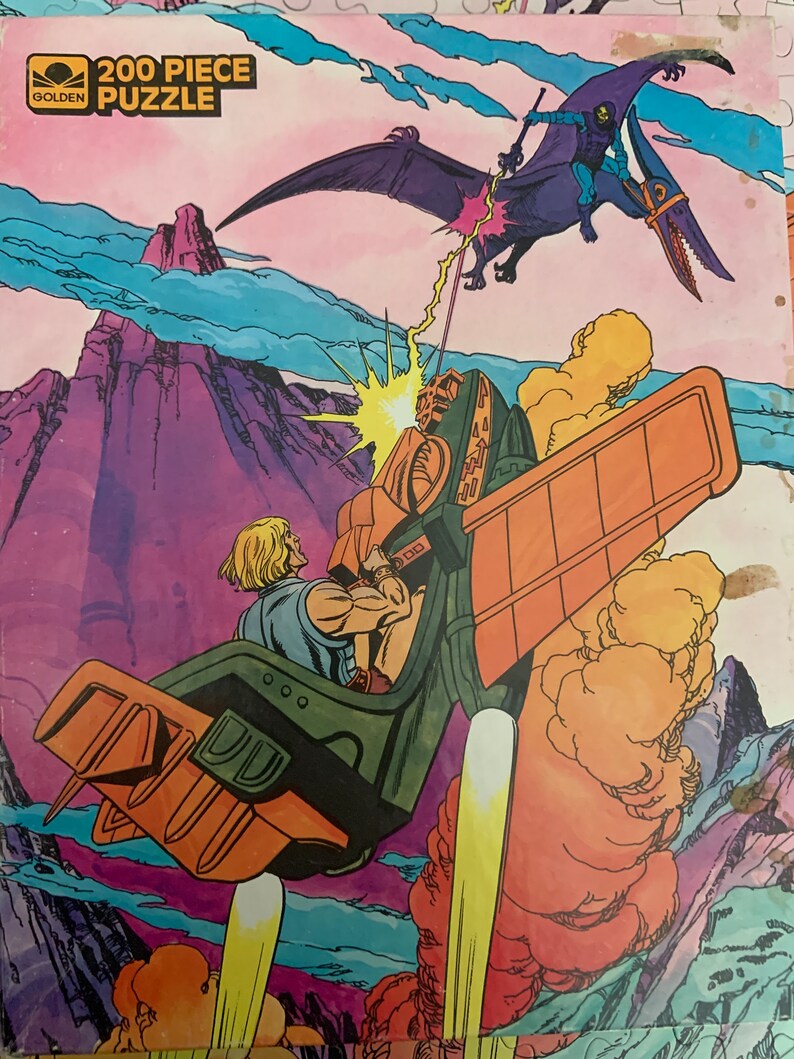 May include: A 200 piece Golden puzzle featuring He-Man flying in his battle-pod, firing a laser beam at a purple flying creature with a sword. The background is a purple and blue sky with a mountain in the distance.