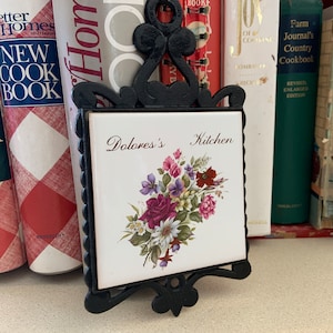Puede incluir: Salvamanteles de hierro fundido negro con una placa de azulejo blanco. El azulejo presenta un ramo floral y el texto "Dolores's Kitchen". El salvamanteles se muestra frente a una colección de libros de cocina.