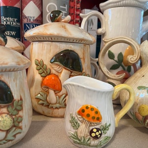 May include: A collection of ceramic mushroom-themed kitchenware. The set includes a sugar bowl, creamer, and pitchers, all decorated with colorful mushroom designs in orange, green, and brown. The pieces have a rustic, whimsical aesthetic.