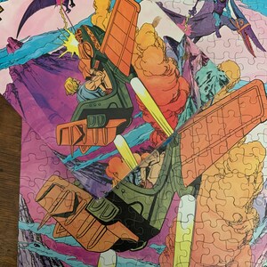 May include: A vintage He-Man and the Masters of the Universe jigsaw puzzle featuring He-Man flying in his battle-pod, Skeletor flying on his winged steed, and a pterodactyl. The puzzle pieces are arranged in a colorful, cartoon-style scene.