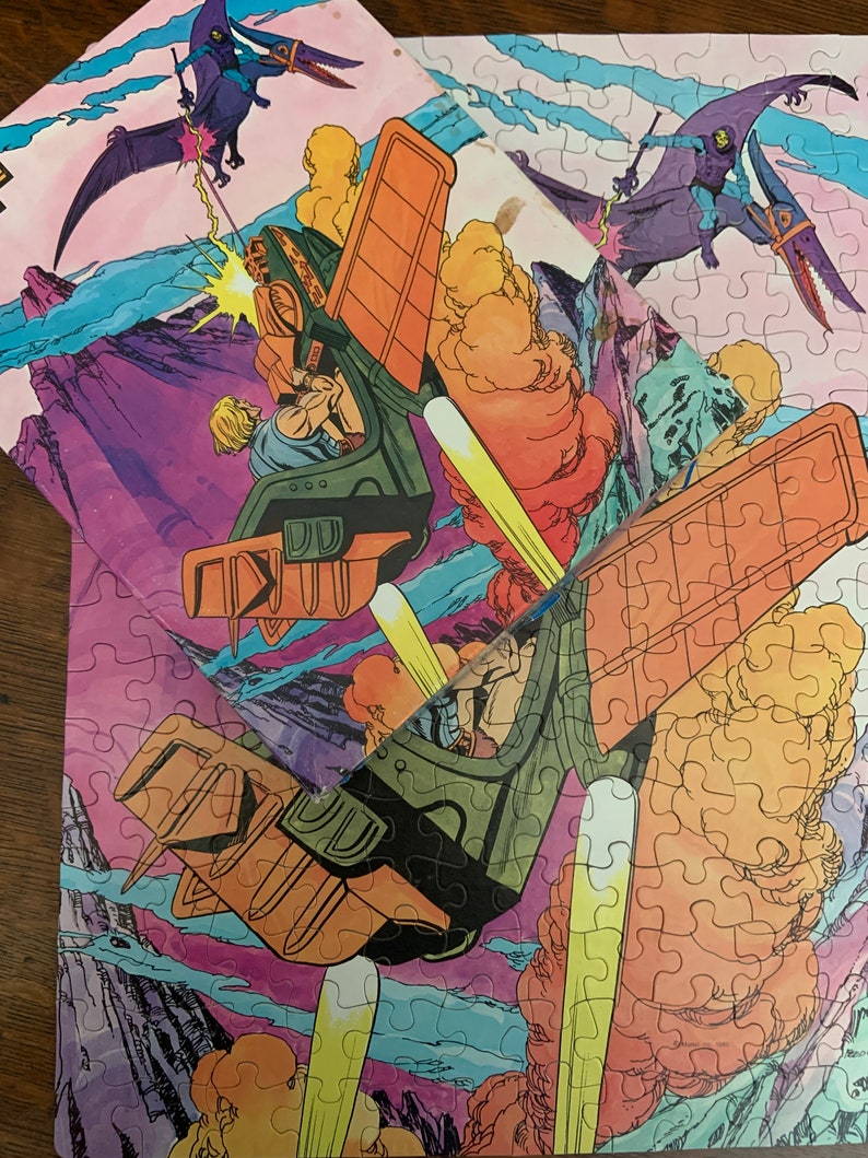 May include: A vintage He-Man and the Masters of the Universe jigsaw puzzle featuring He-Man flying in his battle-pod, Skeletor flying on his winged steed, and a pterodactyl. The puzzle pieces are arranged in a colorful, cartoon-style scene.