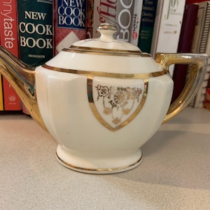 May include: A white teapot with gold trim and floral designs. The teapot has a lid and a handle. The teapot is sitting on a table.