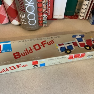 Könnte beinhalten: Eine Vintage-Schachtel mit Tupperware Toys Build-O-Fun Bauklötzen. Die Schachtel zeigt einen roten, weißen und blauen Spielzeugzug und den Text "Build-O-Fun" in großen, fetten Buchstaben. Die Schachtel enthält auch den Text "helle Plastikteile, gleiten zusammen, bewegliche Spielzeuge erstellen, Spaß beim Bauen, Spaß beim Spielen".