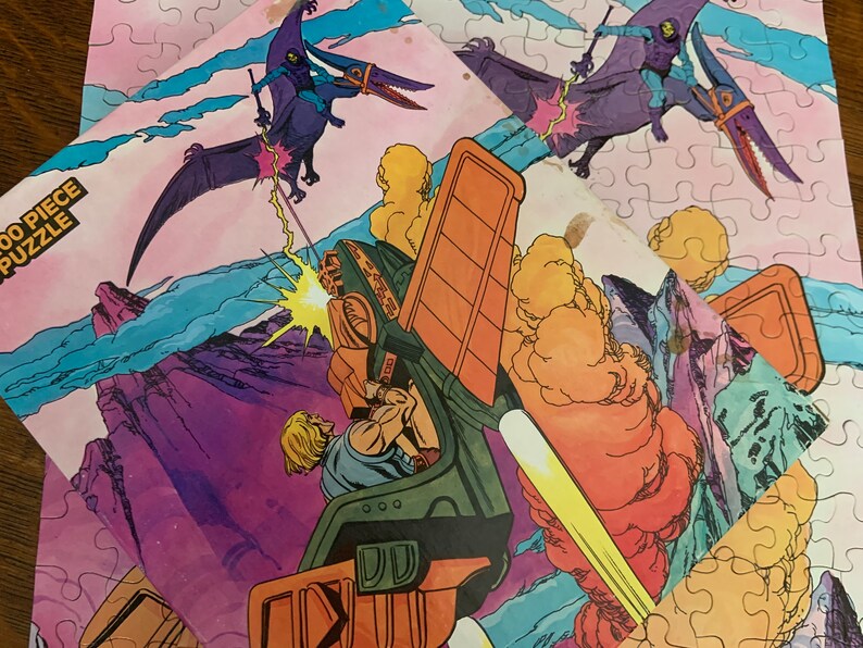 May include: A 100 piece jigsaw puzzle featuring a cartoon illustration of a futuristic vehicle with a purple and green character flying above it. The puzzle is set against a pink, blue, and purple background with clouds and mountains.