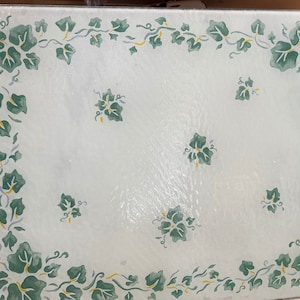 May include: A white glass chopping board with a green ivy design around the edge. The design features green leaves and yellow vines.