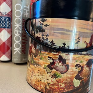 May include: A black metal ice bucket with a hunting scene design. The design features a sunset sky, trees, and three quail birds. The text "The Taste of Home" is printed on the side of the bucket.