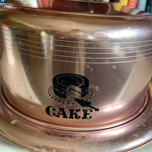 May include: A vintage rose gold cake dome with a black and white cake design and the word "CAKE" printed on the front.