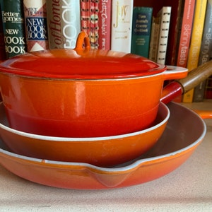 Pode incluir: Um conjunto de utensílios de cozinha esmaltados em laranja, incluindo uma panela com tampa, uma frigideira e uma panela menor. Os utensílios de cozinha têm bordas brancas e alças de madeira. Livros de receitas são visíveis no fundo.