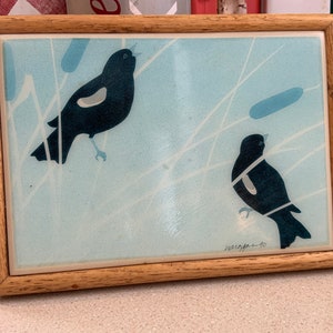 May include: A framed tile featuring a blue background with two black birds, each with a white marking. The birds are perched on branches and the tile is signed "McGeffen-40".