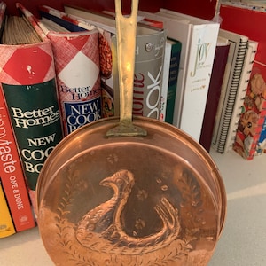 May include: A copper cooking pan with a brass handle. The pan has an embossed design of a bird on the bottom. The pan is sitting on a shelf with cookbooks.