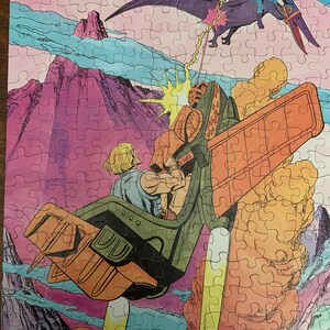 May include: A vintage He-Man puzzle featuring a purple and orange flying creature with a sword, attacking a green and orange spaceship with a man in a blue shirt and yellow pants. The background is a pink and purple mountain range with blue clouds.