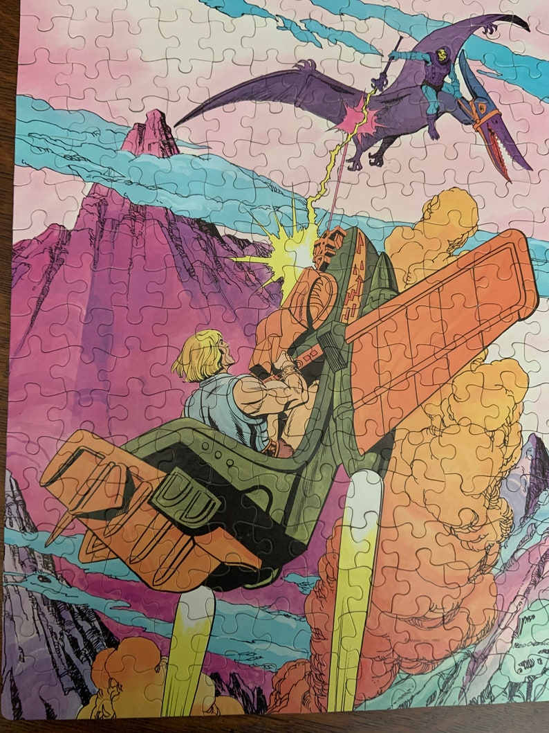 May include: A vintage He-Man puzzle featuring a purple and orange flying creature with a sword, attacking a green and orange spaceship with a man in a blue shirt and yellow pants. The background is a pink and purple mountain range with blue clouds.