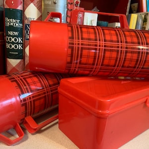 May include: Two red and black plaid thermoses with handles and a red plastic storage box. The thermoses have a black and red plaid pattern. The storage box has a lid.