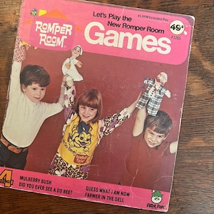 May include: A pink and white record album cover featuring a photo of three children holding puppets. The album is titled "Let's Play the New Romper Room Games" and features the Romper Room logo. The album is priced at 49 cents.