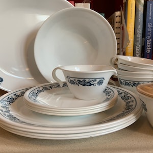 Puede incluir: Un juego de vajilla blanco con un diseño floral azul. El juego incluye platos, cuencos, tazas y una salsera. Los platos están apilados y los cuencos son de varios tamaños. El patrón floral decora los bordes de los platos y las tazas.