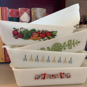 May include: A stack of white rectangular Pyrex baking dishes with various colorful floral and vegetable designs. The dishes are stacked on a shelf, with cookbooks and decorative items in the background.