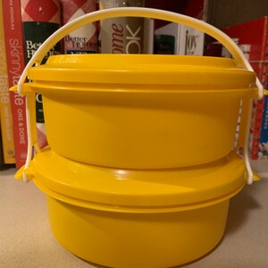 May include: A yellow two-tiered food container with a white handle. The container is made of plastic and has a round shape.