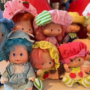 May include: A collection of eight vintage Strawberry Shortcake dolls, each with a unique outfit and hairstyle. The dolls are all dressed in colourful dresses and hats, and some have accessories such as purses or shoes.