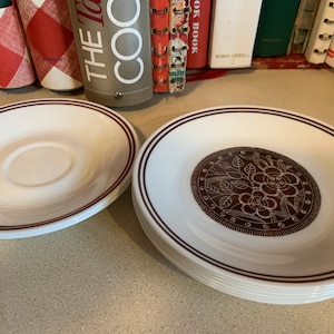 Könnte beinhalten: Zwei Sätze weißer Untertassen aus Keramik mit braunem Rand. Ein Satz hat ein braunes Blumenmuster auf der Oberseite.