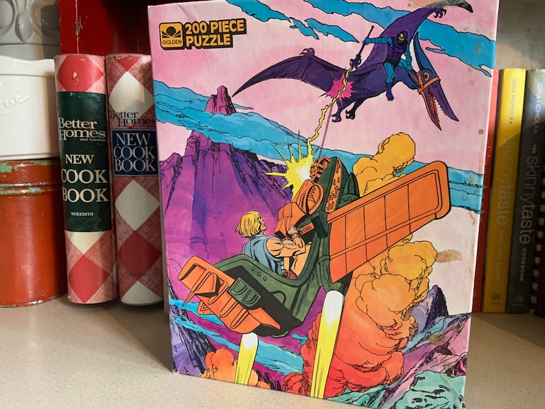 May include: A 200 piece jigsaw puzzle featuring a scene from the Masters of the Universe cartoon.  He-Man is flying in his battle-plane, while Skeletor is flying on a purple dragon.  The background is a pink and blue sky with mountains.