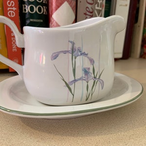 Puede incluir: Salsera de cerámica blanca con borde verde y diseño floral con iris morados. La salsera está sobre una base a juego.