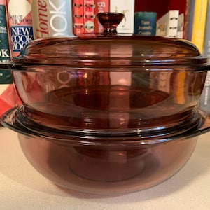 May include: A set of two clear glass casserole dishes with lids. The dishes are a deep purple colour and have a round shape. The lids are also clear glass and have a knob on top.