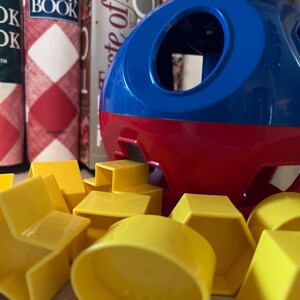 Puede incluir: Un juguete clasificador de formas rojo y azul con varias formas geométricas amarillas. El juguete tiene forma esférica con recortes para las formas. Las formas incluyen una estrella, un círculo, un cuadrado y un hexágono. Libros de cocina en el fondo.