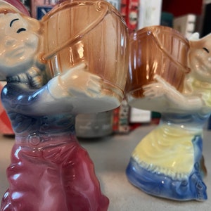 Puede incluir: Un par de figuras de cerámica vintage que representan figuras sonrientes que llevan cubos de madera. Las figuras visten ropa colorida en tonos azules, amarillos y rojos. Las figuras son probablemente saleros y pimenteros.