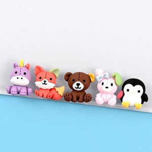 May include: Five colorful, small, cartoon animal figurines. A purple unicorn, an orange fox, a brown bear, a pink bunny rabbit, and a black penguin.