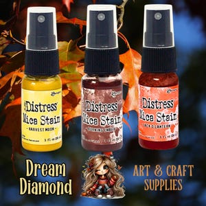 Puede incluir: Tres botellas de Distress Mica Stain en amarillo, marrón y naranja, cada una con una boquilla pulverizadora negra. Las botellas están etiquetadas como "Harvest Moon", "Burning Ember" y "Jack O'Lantern". La imagen también incluye el texto "Dream Diamond" y "ART & CRAFT SUPPLIES".