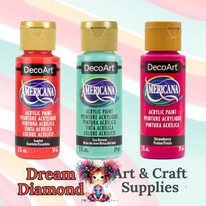 May include: Three bottles of DecoArt Americana acrylic paint in red, sea green, and pink. Each bottle has a gold cap and contains 59 ml of paint. The labels include the text "Acrylic Paint" and colour names like "Scarlet" and "Strawberry."