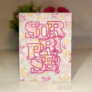 May include: A pink and white card with a floral pattern and the word "SURPRISE" in pink letters with a white outline. The card is designed by Karen McKibbin Creative Expressions.