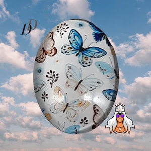 May include: A white oval-shaped cabochon with a butterfly design. The butterflies are blue, brown, and white with a white background.