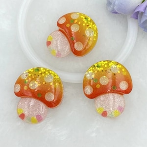 May include: Three orange and yellow mushroom shaped resin cabochons with white and yellow glitter accents.
