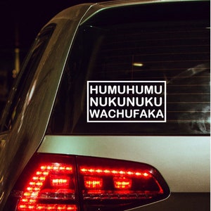 May include: A white rectangular sticker on a car's rear window. The sticker has the words "HUMUHUMU NUKUNUKU WACHUFAKA" in a bold, sans-serif font. The car's taillights are illuminated, and the background is dark.